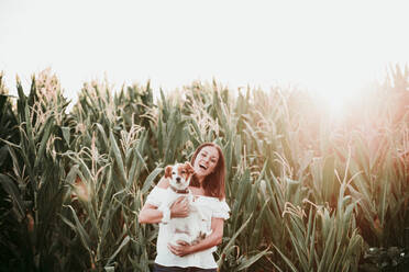 Happy woman with dog at cornfield during sunset - EBBF00671