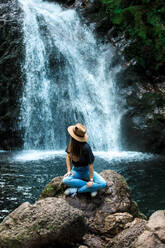 Fröhliche Reisende mit Hut lächelt und schaut in die Kamera, während sie auf einem nassen Felsen in der Nähe eines Wasserfalls sitzt - ADSF14391