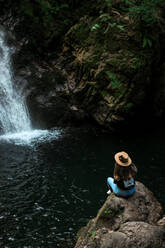 Cheerful female traveler in hat smiling and looking at camera while sitting on wet boulder near waterfall - ADSF14393