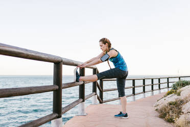 Seitenansicht einer sportlichen Frau, die sich beim Training auf der Promenade am Meer aufwärmt und die Beine dehnt - ADSF14397
