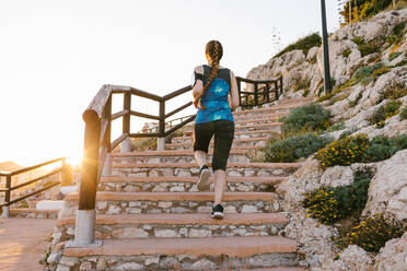 Niedriger Blickwinkel von hinten auf eine nicht erkennbare Sportlerin in T-Shirt und Leggings, die beim Training während des Sonnenuntergangs die Steinstufen hinaufläuft - ADSF14399