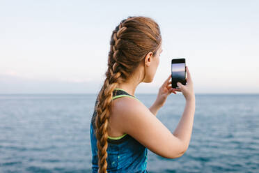 Seitenansicht einer jungen Frau, die auf einer Böschung steht und mit ihrem Smartphone ein Foto von der beeindruckenden Meereslandschaft macht - ADSF14400