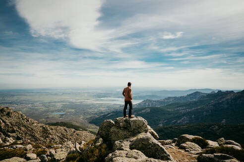Rückenansicht eines anonymen Mannes mit Händen in den Taschen, der auf einem Stein steht und die Bergkette Puerto de la Morcuera an einem bewölkten Tag in Spanien bewundert - ADSF14415