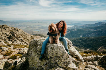 Voller Länge weiblichen Besitzer umarmt flauschigen Labradoodle beim Sitzen auf Stein gegen bewölkten Himmel in Puerto de la Morcuera Berge in Spanien - ADSF14418