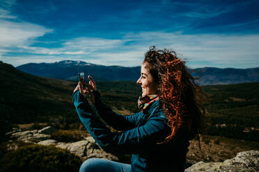 Side view of glad woman using smartphone to take photos of Puerto de la Morcuera mountain ridge on cloudy day in Spain - ADSF14420