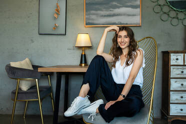 Pleasant young female in trendy outfit looking at camera with smile while sitting in stylish design armchair in room with modern interior - ADSF14424