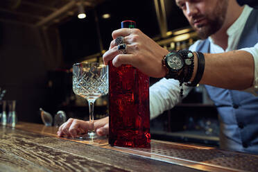 Cropped professional young bartender holding red bottle while preparing a cocktail in the bar - ADSF14427