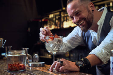 Fröhlicher professioneller Barkeeper, der in der Bar einen Cocktail mit getrockneten Erdbeeren dekoriert - ADSF14430