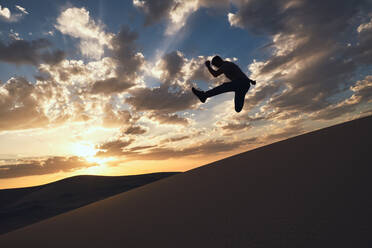 Silhouette eines anonymen fitten männlichen Tänzers, der einen Trick mit ausgestrecktem Bein über sandigem Gelände in der Wüste bei hellem Sonnenuntergang in der Nacht vorführt - ADSF14446