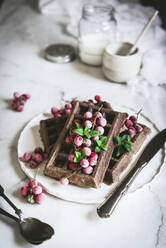 Von oben leckere frische Haferflocken-Schokowaffeln mit Stachelbeeren auf weißem Teller mit Löffel und Messer auf hellem Holztisch serviert - ADSF14449