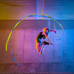 Woman jumping while spinning ribbon against wall - STSF02598