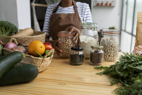 Woman with apron storing pulses in jars on table with fresh fruit and vegetables - VABF03359