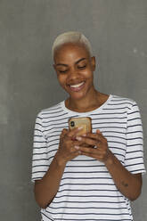 Smiling woman with striped shirt standing in front of gray background, holding smartphone - VABF03409