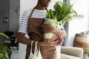 Pretty woman in apron carrying net with fresh vegetables - VABF03414