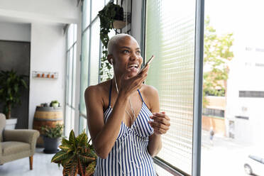 Woman standing at home using smartphone and headphones - VABF03450