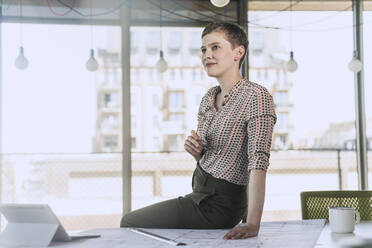 Confident businesswoman sitting on desk in office - UUF21097