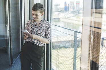 Smiling businesswoman using smartphone at the window in office - UUF21099