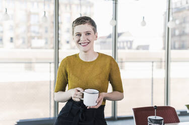 Porträt einer lächelnden Geschäftsfrau im Büro, die einen Kaffeebecher hält - UUF21123