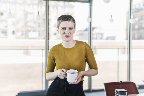 Porträt einer lächelnden Geschäftsfrau im Büro, die einen Kaffeebecher hält - UUF21124