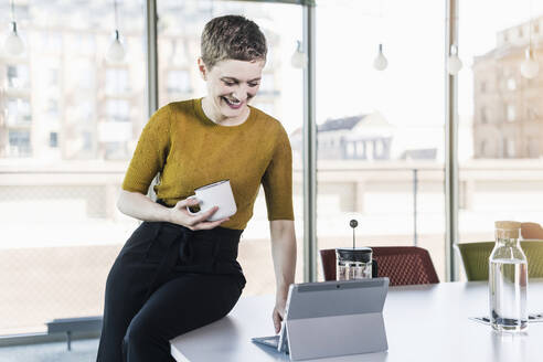 Lächelnde Geschäftsfrau sitzt auf einem Schreibtisch im Büro, hält einen Kaffeebecher und benutzt ein Tablet - UUF21126