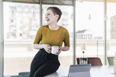 Laughing businesswoman sitting on desk in office holding coffee mug - UUF21128