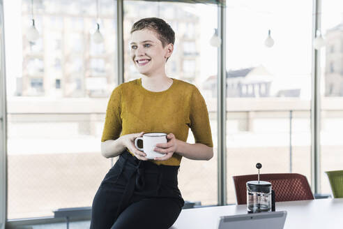 Lächelnde Geschäftsfrau sitzt auf einem Schreibtisch im Büro und hält einen Kaffeebecher - UUF21129