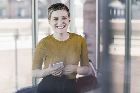 Porträt einer lächelnden Geschäftsfrau im Büro, die ein Mobiltelefon hält - UUF21131