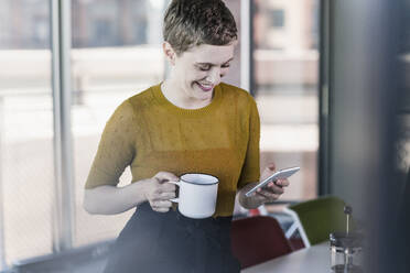 Smiling businesswoman in office holding coffee mug and mobile phone - UUF21134