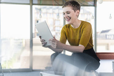 Happy businesswoman sitting on desk in office using tablet - UUF21146