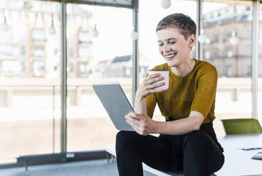 Businesswoman sitting on desk in office using tablet - UUF21148