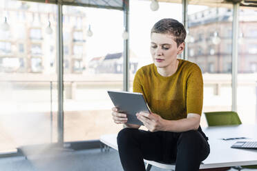 Businesswoman sitting on desk in office using tablet - UUF21149