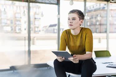 Thoughtful businesswoman sitting on desk in office holding tablet - UUF21150