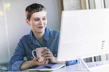 Lächelnde Geschäftsfrau am Schreibtisch im Büro mit Blick auf den Computerbildschirm - UUF21166