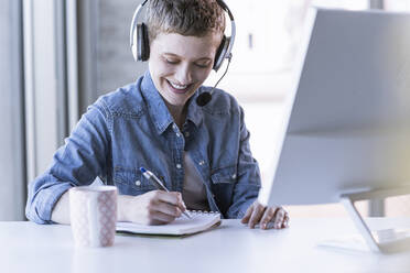 Smiling businesswoman wearing headset at desk in office taking notes - UUF21180