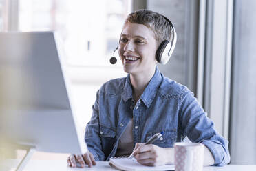 Lächelnde Geschäftsfrau mit Headset am Schreibtisch im Büro, die Notizen macht - UUF21181