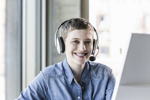 Portrait of smiling businesswoman wearing headset at desk in office - UUF21184