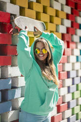 Woman with hand raised holding skateboard while standing against colored wall - DLTSF01119