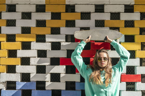 Junge Frau, die ein Skateboard anhebt, während sie gegen eine Backsteinmauer steht, lizenzfreies Stockfoto