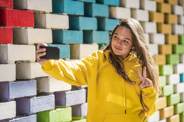 Young woman taking selfie on smart while standing by colored brick wall - DLTSF01125