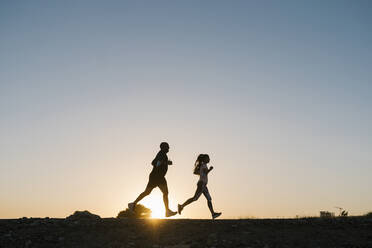 Silhouette eines Sportlers und einer Frau, die gegen den klaren Himmel bei Sonnenuntergang laufen - EGAF00691