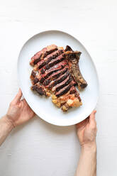 From above crop hands of person holding rib steak served with potato on white background - ADSF14466