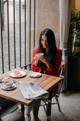 From above side view of modern married couple sitting at small wooden table with newspaper and having breakfast and drinking coffee in cafe - ADSF14468