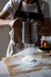 Crop person in apron standing by table sifting flour on baking paper with stainless sifter for making dough - ADSF14490