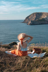 Back view of woman in hat with glass of drink sitting on mat for picnic on dry shore and romantically looking along to serene water and hills - ADSF14494