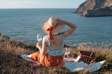Rückenansicht einer Frau mit Hut und einem Glas Getränk, die auf einer Picknickmatte am trockenen Ufer sitzt und romantisch auf das ruhige Wasser und die Hügel blickt - ADSF14495