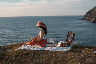 Seitenansicht einer Frau mit Hut und Buch, die auf einer Matte sitzt und ein Picknick mit Wassermelone und einem Glas Getränk am trockenen Ufer macht und romantisch auf das ruhige Wasser blickt - ADSF14500