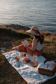Seitenansicht einer Frau mit Hut und Buch, die auf einer Matte sitzt und ein Picknick mit Wassermelone und einem Glas Getränk am trockenen Ufer macht und romantisch auf das ruhige Wasser blickt - ADSF14501