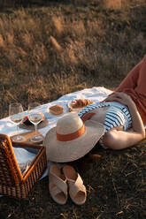 Von oben genießt Frau auf weißer Picknickmatte liegend mit Hut im Gesicht in der Nähe von Korb auf Rasen - ADSF14502