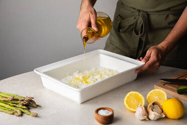 Anonymer Koch gießt Olivenöl in ein Backblech mit gehackten Zwiebeln, während er das Abendessen am Küchentisch vorbereitet - ADSF14518