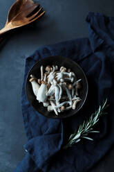Draufsicht auf eine Schüssel mit ungekochten Pilzen und frischen grünen Rosmarinstängeln auf einem dunkelblauen Handtuch in einem Arrangement mit Holzlöffel und Gabel auf einem schwarzen Tisch in einer rustikalen Küche - ADSF14531